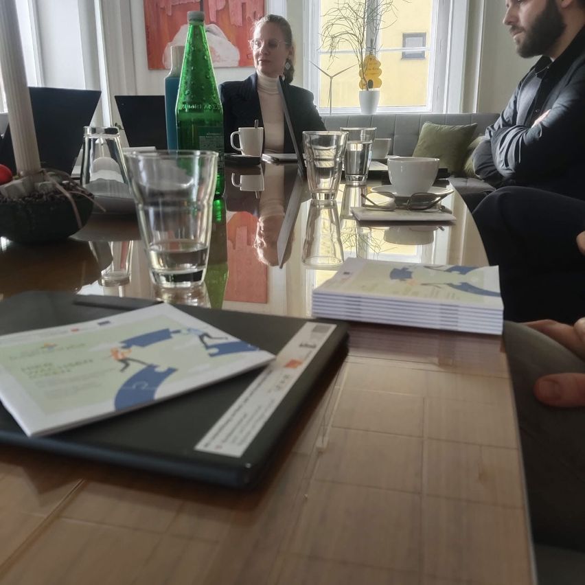 A man and a woman sit at a table in a meeting room, both wearing business attire. The table is set with cups, glasses, and a green bottle. There are two laptops on the table.