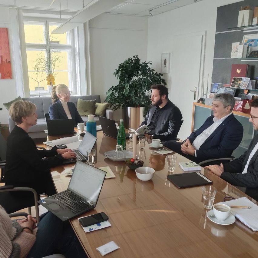 Six individuals, both men and women, are engaged in a meeting inside a conference room. Laptops, books, and cups are on the table. There is a plant and picture frames on the wall.