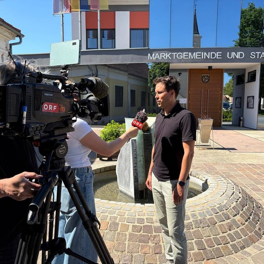 Ein Mann wird von einer Reporterin vor einem Gebäude mit Fahnen interviewt. Der Mann hält ein Mikrofon und trägt eine Uhr. Hinter ihm steht eine Kamera auf einem Stativ.