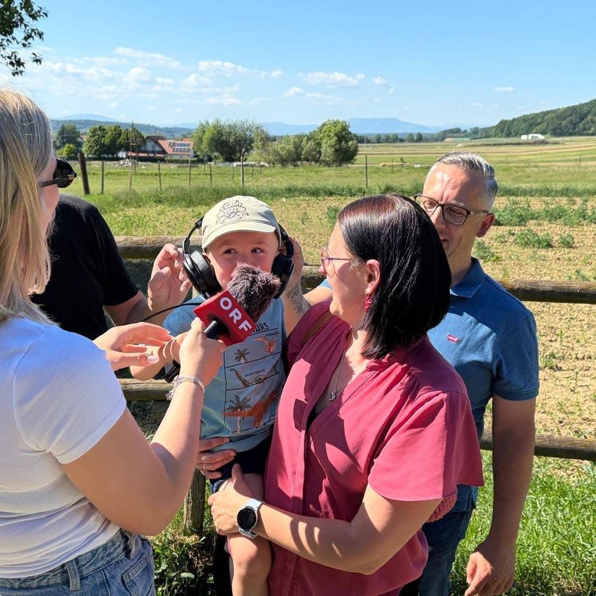 Eine Frau interviewt einen kleinen Jungen mit Kopfhörern auf einem Feld, während ein Mann und eine andere Frau zusehen.