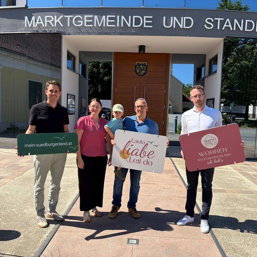 Vier Personen stehen vor einem Gebäudeeingang und halten Schilder in der Hand. Eine Frau und ein Kind stehen in der Mitte. Das Gebäude ist mit 'Marktgemeinde und Stand' beschriftet.