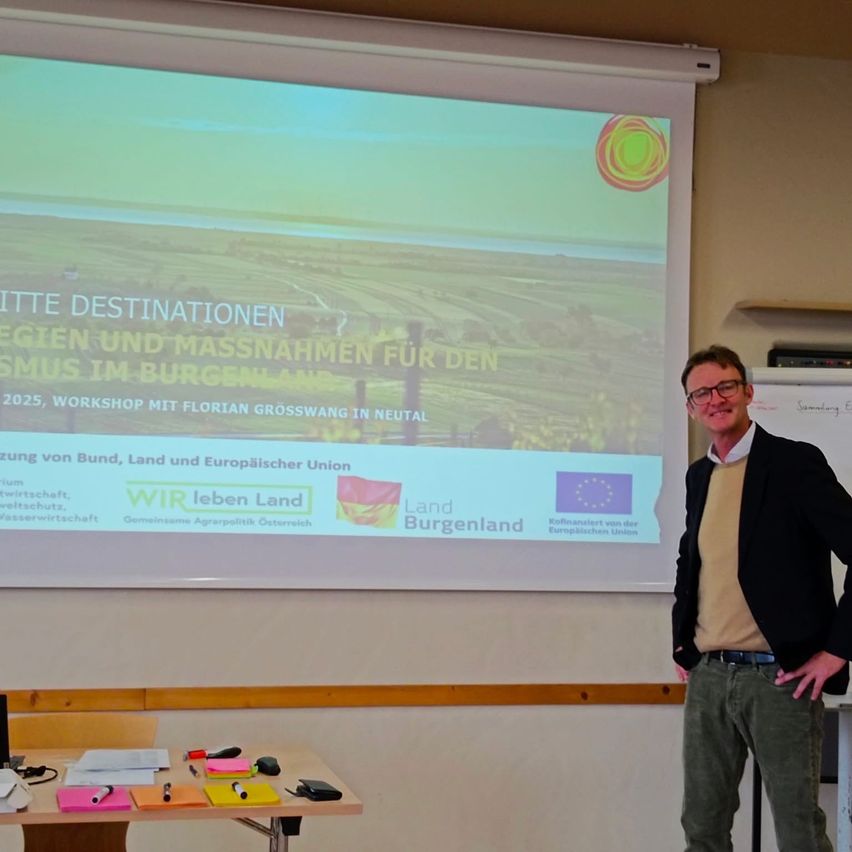 Ein Mann steht vor einer Projektionsleinwand mit einem Workshop-Titel auf Deutsch. Er trägt einen Anzug und eine Brille.