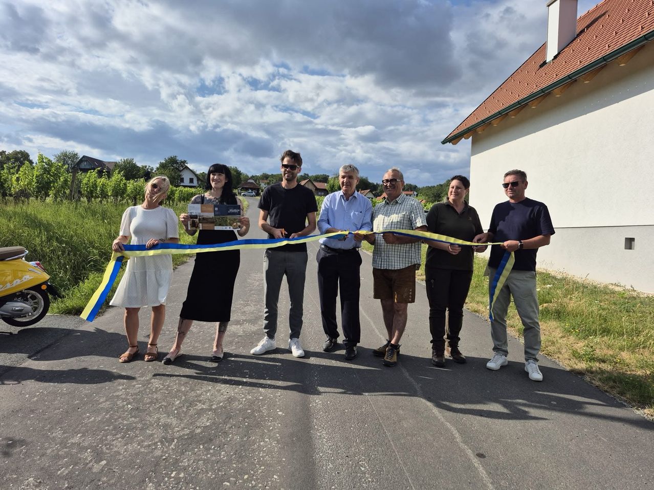 Eine Gruppe von Menschen steht auf einer Straße und hält ein blau-gelbes Band, wahrscheinlich zur Feier einer Eröffnung.