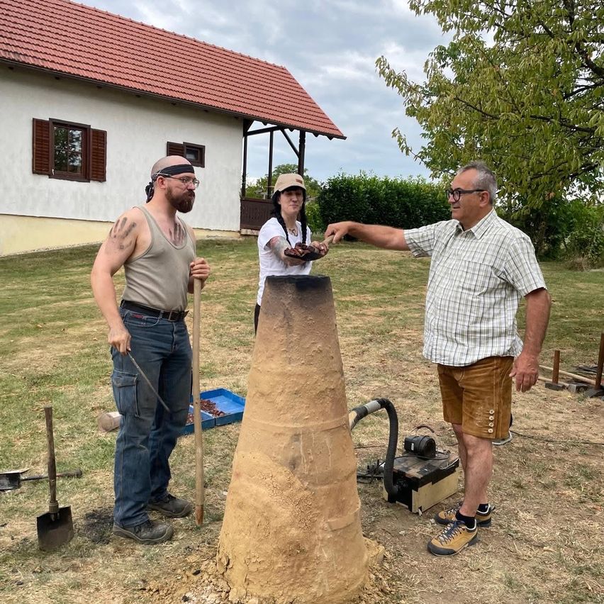 Drei Personen stehen vor einem Haus. Ein Mann zeigt auf eine große Tonstruktur, ein anderer Mann hält eine Schaufel, und eine Frau steht in der Nähe.