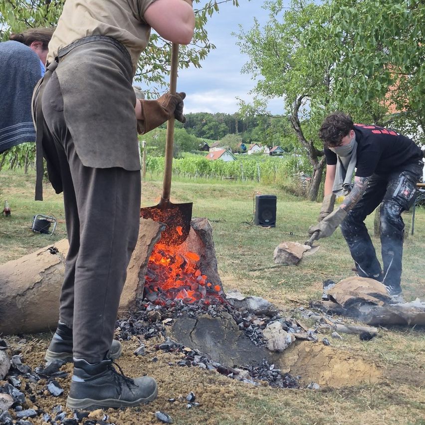 Zwei Männer arbeiten an einem Feuer im Freien, einer hält eine Schaufel und der andere einen Stein. Sie sind von Steinen und einem Feuer umgeben.