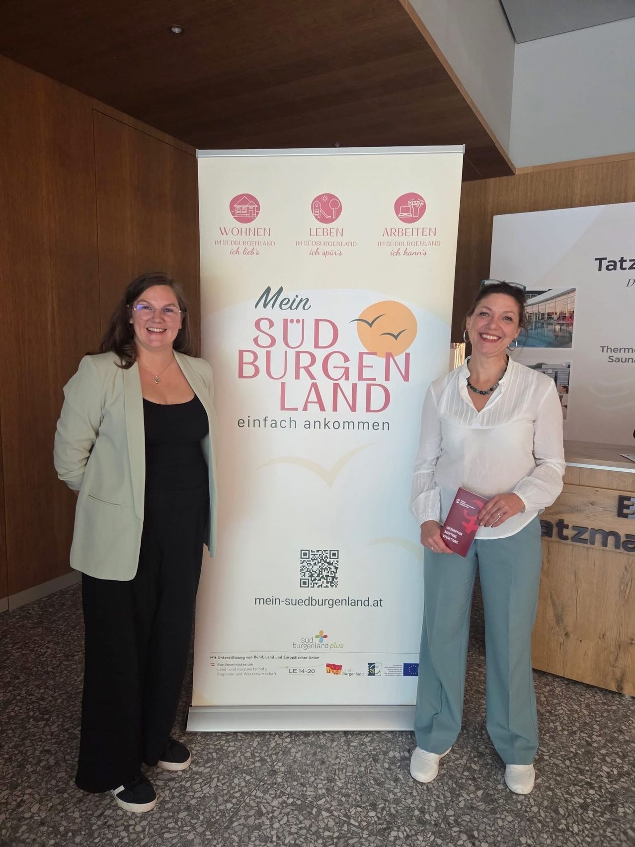 Zwei Frauen stehen neben einem Werbebanner für 'Mein Süd Burgenland'. Das Banner zeigt Symbole für Wohnen, Freizeit und Arbeit. Die Frau links trägt eine helle Jacke und eine Brille, während die Frau rechts ein Buch in der Hand hält und ein weißes Oberteil trägt. Hinter ihnen befindet sich eine Holztheke mit einem Schild.