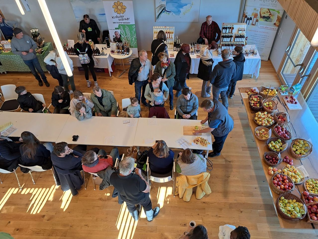 An overhead view of a room with people sitting and standing around tables, some are looking at products on the tables, others are talking. There are baskets of apples on the side.