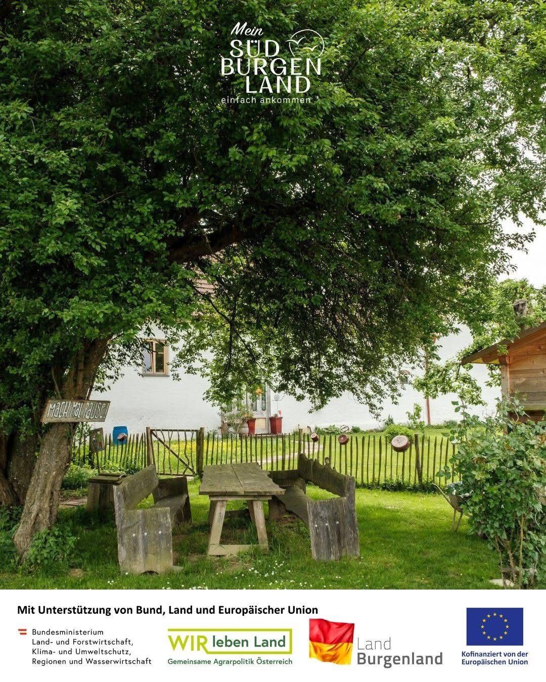 Eine ruhige Gartenszene mit einer Holzbank, einem Tisch und einem Schild unter einem großen Baum. Hinter der Bank ein weißes Haus mit Zaun und Schubkarren.