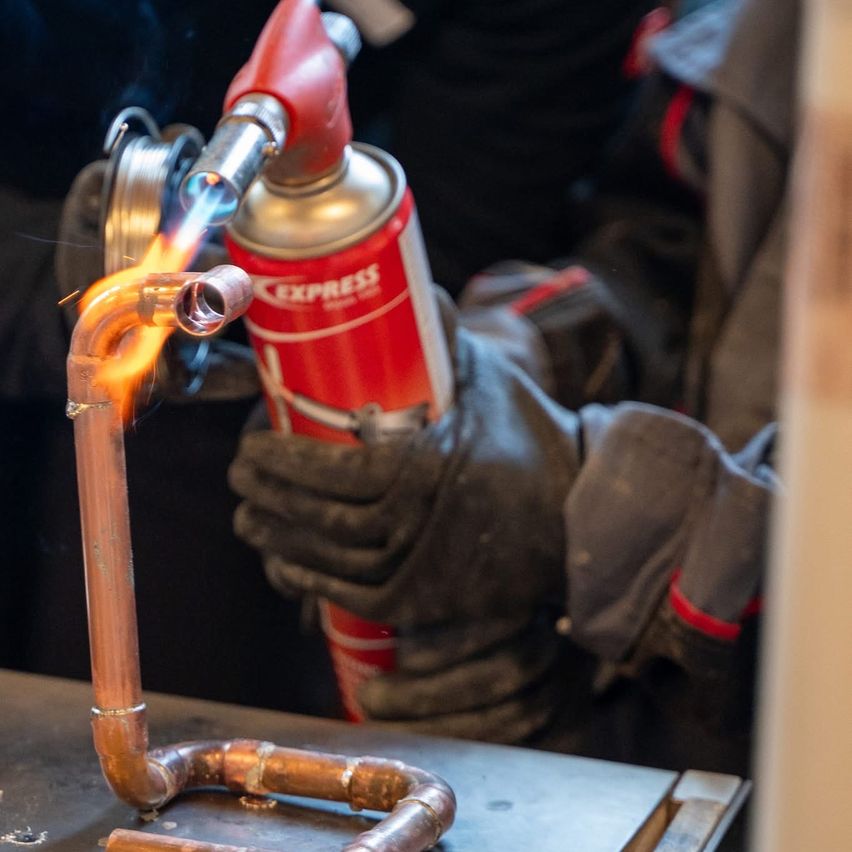 Eine Person mit Handschuhen lötet ein Kupferrohr mit einem Brenner. Der Brenner gibt eine Flamme ab und das Kupferrohr wird erhitzt.