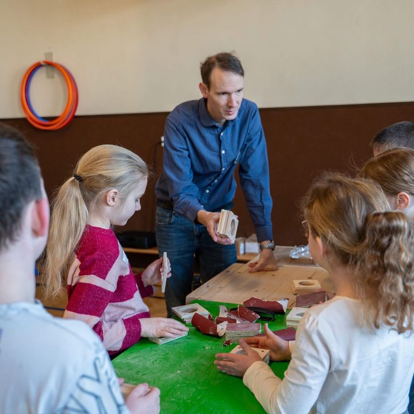 Ein Mann in einem blauen Hemd unterrichtet eine Gruppe von Kindern bei einem Bastelprojekt an einem Tisch. Die Kinder sitzen um den Tisch herum, und eine hält ein kleines Objekt.
