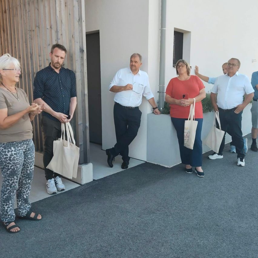 Eine Gruppe von Menschen, darunter zwei Männer und drei Frauen, steht vor einem Gebäude und hält weiße Taschen. Sie sind lässig gekleidet und scheinen ein Gespräch zu führen.