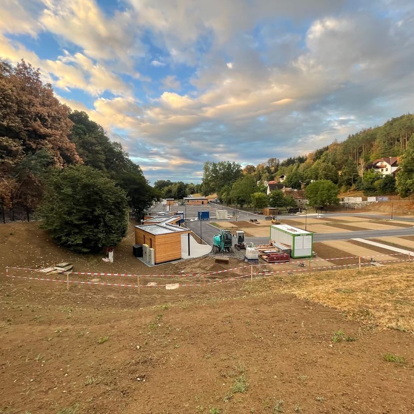 A construction site with a building under construction, surrounded by trees and a road. Equipment and materials are scattered around.