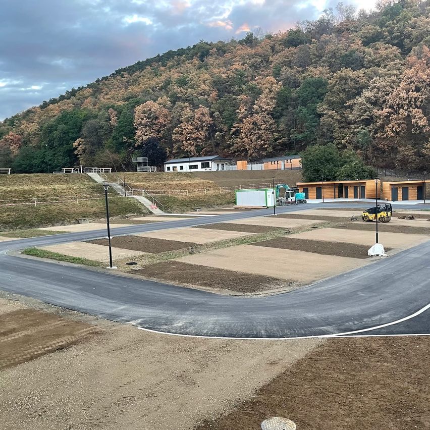 A construction site with a newly paved road, surrounded by a hillside with autumn trees and a few buildings. A yellow machinery vehicle is parked on the right.