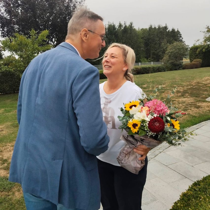 A man and a woman stand together on a pathway, the woman holding a bouquet of flowers and smiling.