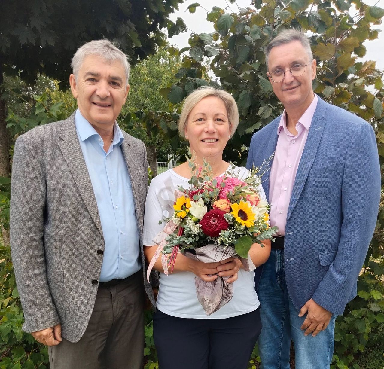 Three people stand together in a garden, smiling. The woman in the middle holds a bouquet of flowers. The man on the left wears a gray jacket, the man on the right wears a blue blazer, and the woman wears a white shirt.