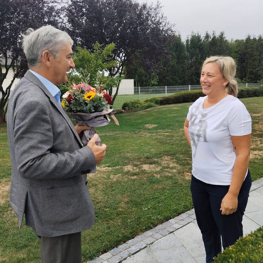 A man in a gray suit holds a bouquet of flowers and talks to a smiling woman in a white shirt in a park.