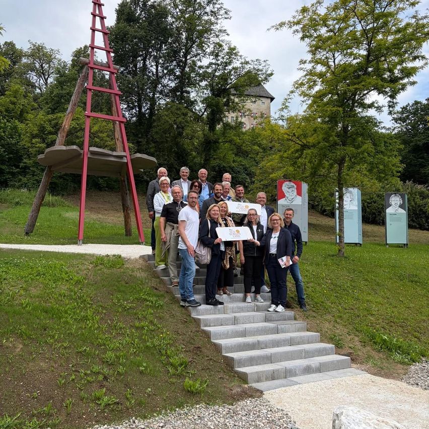 A group of people stands on stairs in a park, holding a certificate. Behind them is a large red structure and a building.