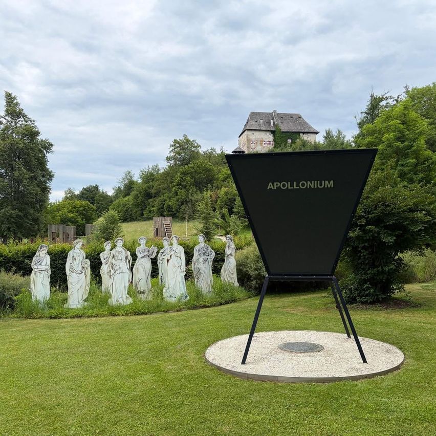 A grassy area has a black sign with Apollonium written on it. Nine statues of women are in a line. There is a castle in the background.