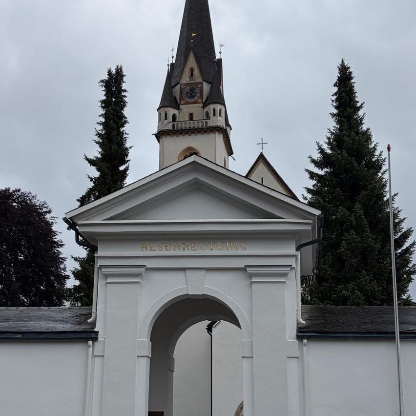A white building with a tower and arched entrance, labeled 'Resurrectus Est.' Trees and a flagpole are in the background.