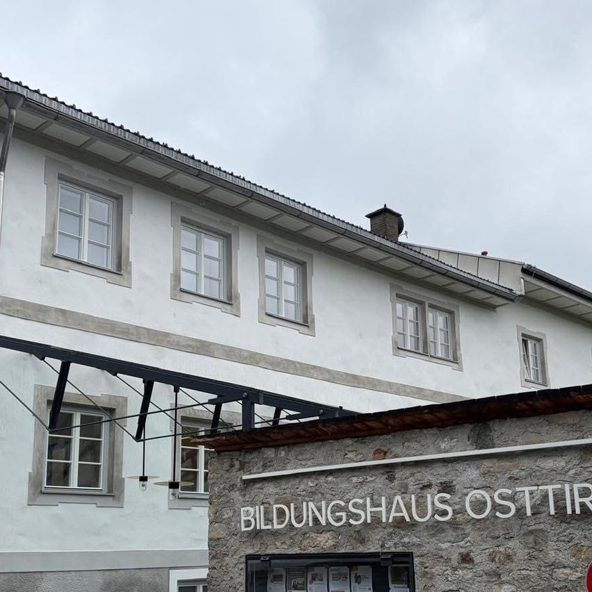 A white building with multiple windows and a brick wall with the text 'BILDUNGSHAUS OSTIIR'. The sky is cloudy.