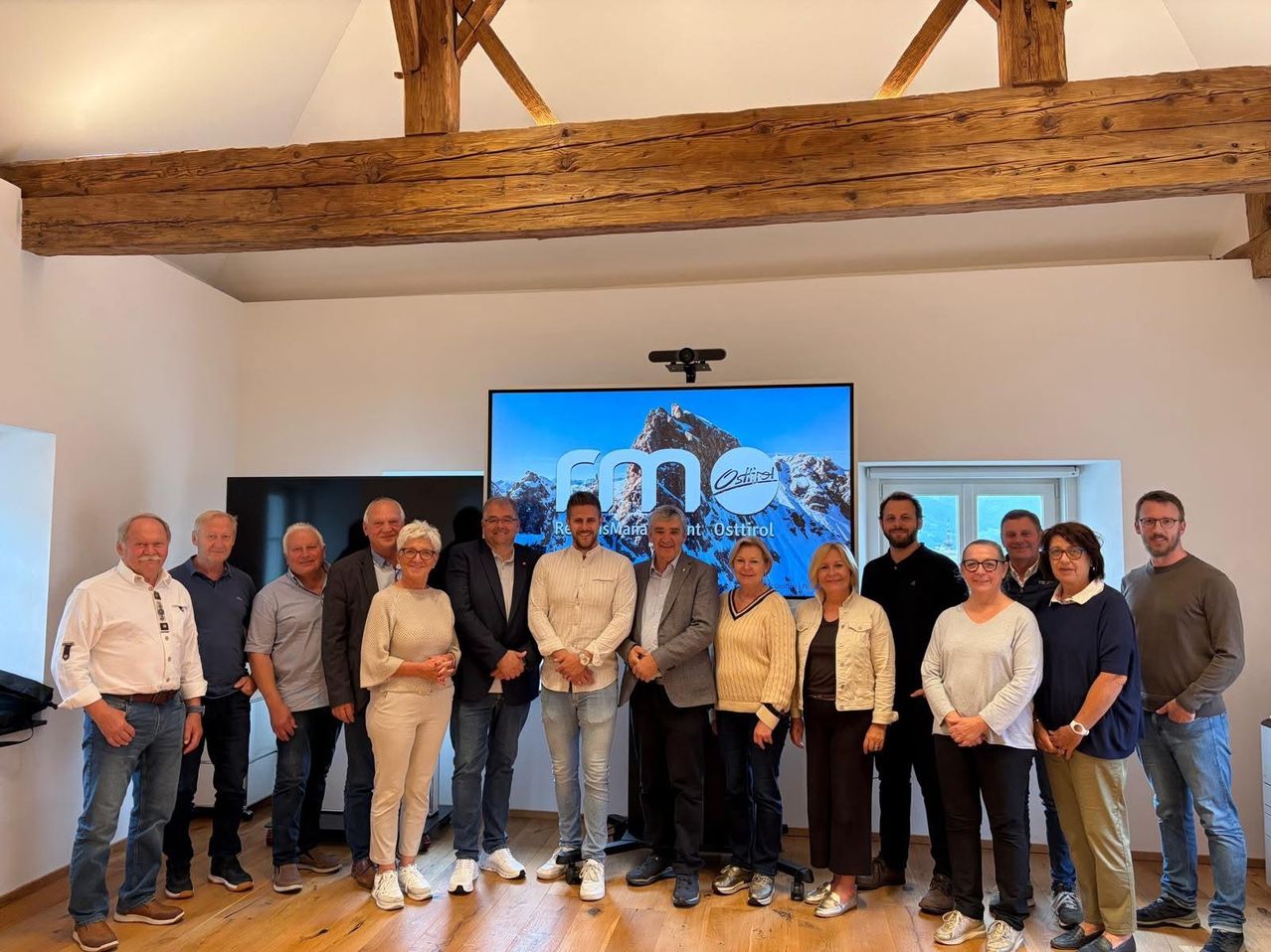 A group of people in formal wear pose for a photo in a room with a large screen displaying mountains. The room has wooden floors and wooden beams on the ceiling.