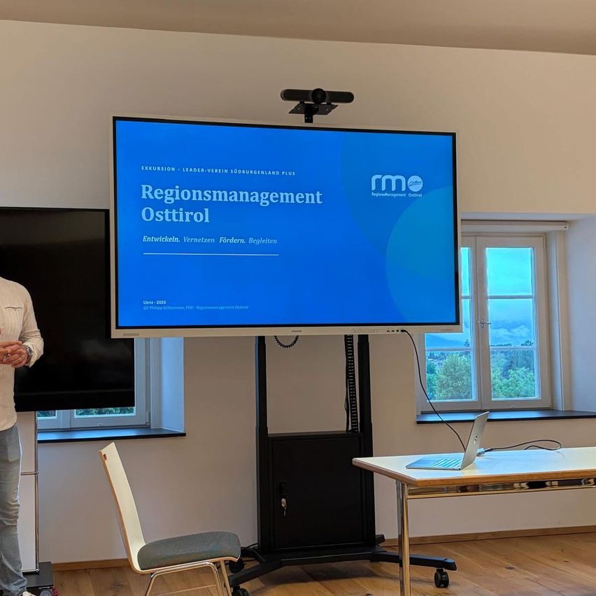 A room with a presentation screen displays 'Regionsmanagement Osttirol'. A man stands nearby, and a laptop sits on a table with a window behind it.