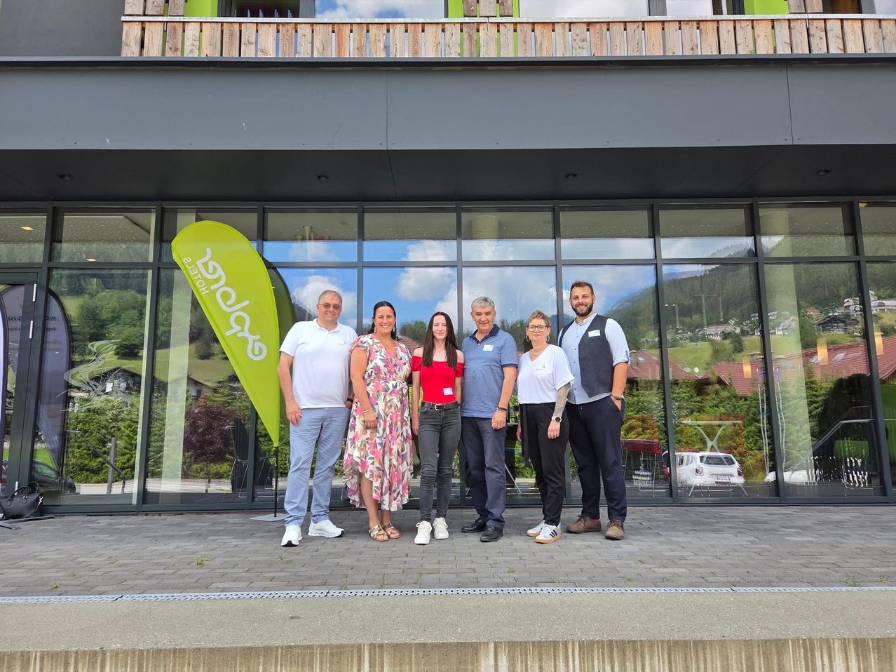 Eine Gruppe von sechs Personen steht vor einem Gebäude mit Glaswänden und lächelt für ein Foto. Sie tragen Freizeitkleidung, wobei eine Frau ein rotes Shirt und eine andere ein Blumenkleid trägt. Hinter ihnen zeigt ein grüner Banner weißen Text, und ein Auto ist durch das Glas zu sehen.
