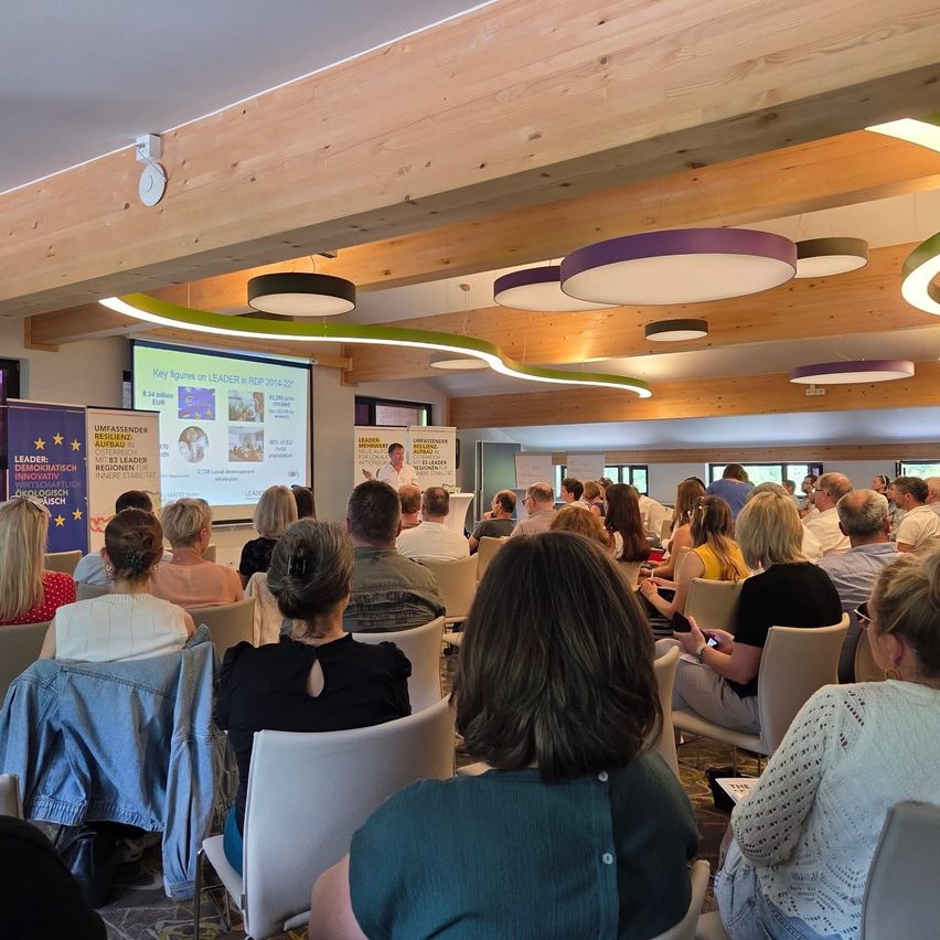 Eine Gruppe von Menschen nimmt in einem Konferenzraum mit Holzdecken an einer Präsentation teil. Eine Beamerleinwand zeigt Schlüsselzahlen, und ein Mann spricht auf der Bühne.