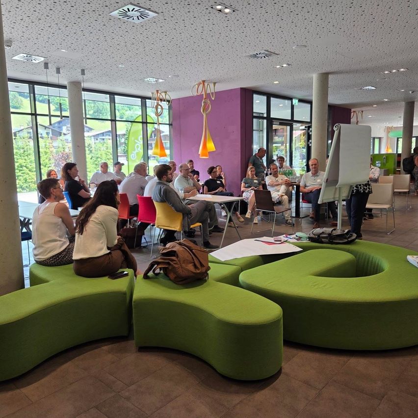 Eine Gruppe von Menschen sitzt um Tische in einem modernen Büroraum. Eine Frau steht bei einer Tafel. Grüne bohnenförmige Sitzgelegenheiten und dekorative Beleuchtung sind vorhanden.