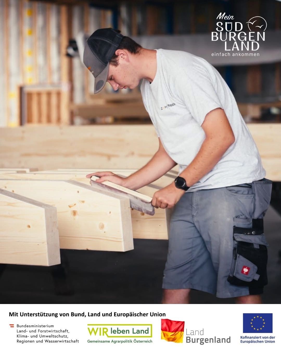 Ein Mann arbeitet mit einer Säge an Holzbalken, trägt ein graues Hemd und eine Kappe, in einer Werkstatt. Hinter ihm sind Holzstrukturen zu sehen und ein Logo für 'Burgen Land' ist oben angebracht.