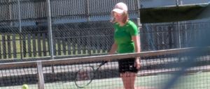 Eine Frau in einem grünen Shirt und einer weißen Kappe steht auf einem Tennisplatz und hält eine Tennisschläger. Ein Tennisball ist in der Luft, und ein Zaun mit einem Netz ist hinter ihr.