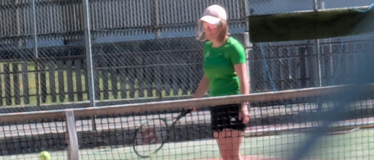 Eine Frau in einem grünen Shirt und einer weißen Kappe steht auf einem Tennisplatz und hält eine Tennisschläger. Ein Tennisball ist in der Luft, und ein Zaun mit einem Netz ist hinter ihr.