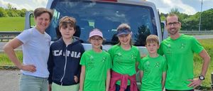Eine Familie mit fünf Personen steht vor einem weißen Van, mit einem bewölkten Himmel im Hintergrund. Sie tragen alle grüne Shirts und lächeln.