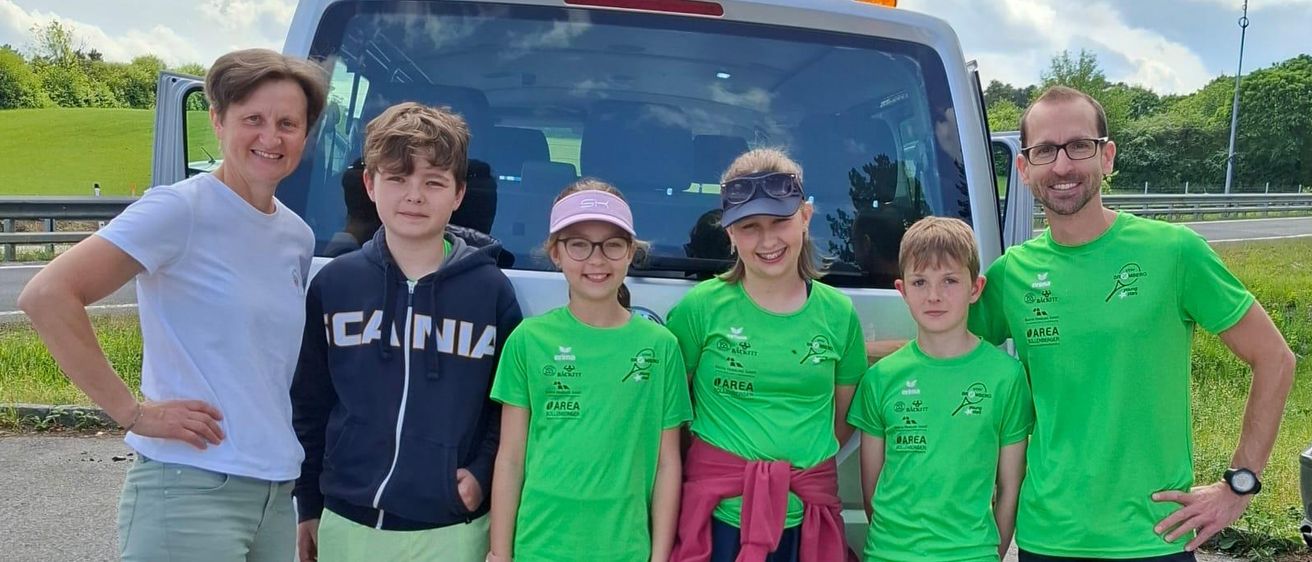 Eine Familie mit fünf Personen steht vor einem weißen Van, mit einem bewölkten Himmel im Hintergrund. Sie tragen alle grüne Shirts und lächeln.
