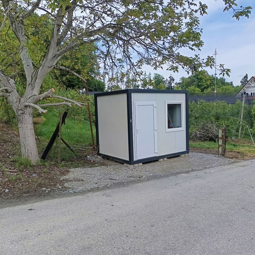 A small, white portable building with a door and window is situated on the side of the road near a tree.