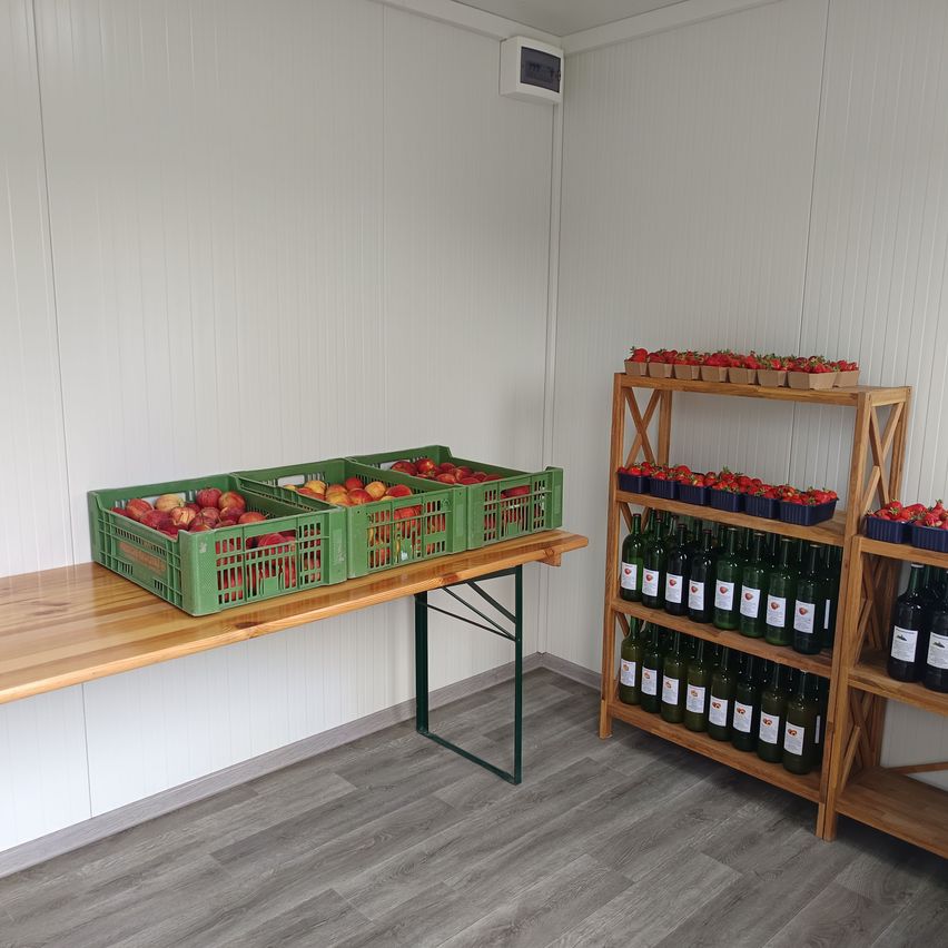 A room with white walls and wooden shelves. Three green crates of peaches sit on a table. Bottles of wine are displayed on a shelf to the right.