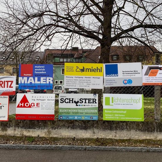 Verschiedene Schilder sind entlang eines Zauns angebracht, darunter solche für Maler Anstrich Fassaden, MIEHL und LeoGRAF. Ein Baum und Gebäude sind im Hintergrund zu sehen.