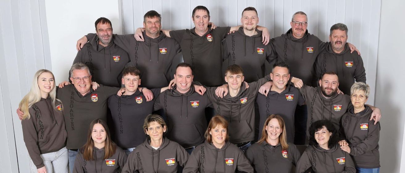 Eine Gruppe von Menschen in schwarzen Jacken mit weißen Schriftzügen posiert für ein Foto. Sie stehen und sitzen auf Stühlen in einem Raum.