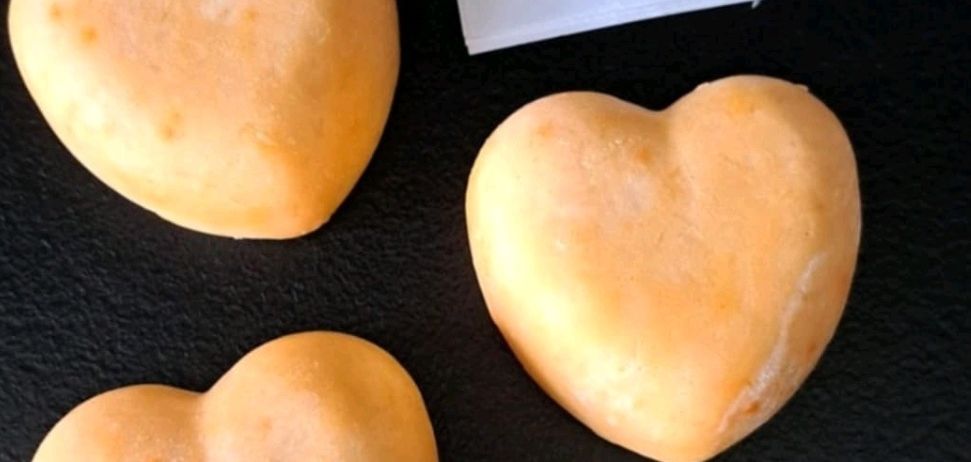 Drei herzförmige Objekte mit einem Etikett, das 'Duschbutter Pfirsich' liest, auf einer schwarzen Oberfläche. Die Objekte scheinen Seife oder Badebälle zu sein.