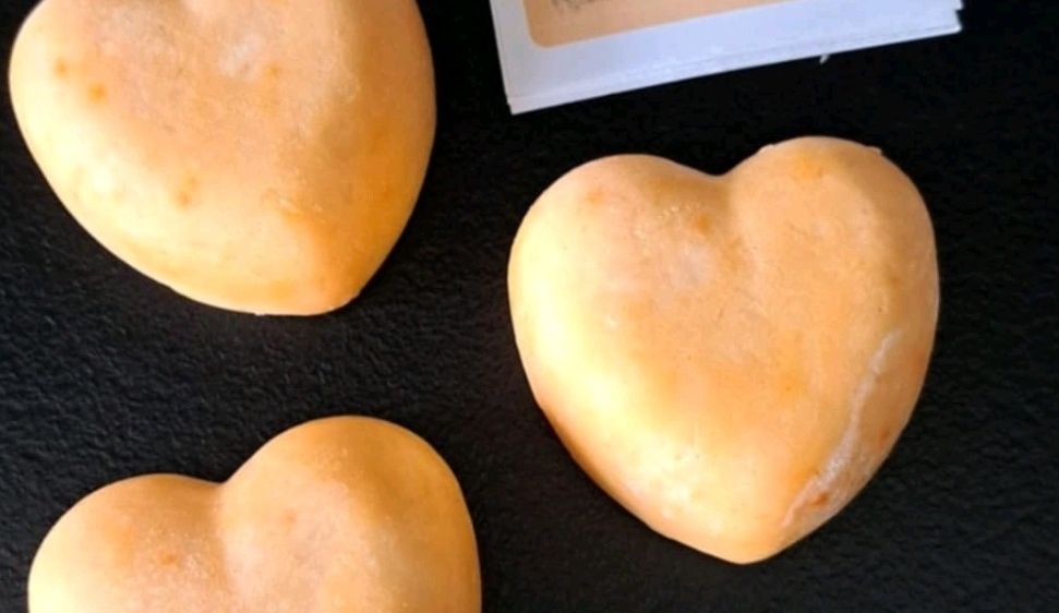 Drei herzförmige Objekte mit einem Etikett, das 'Duschbutter Pfirsich' liest, auf einer schwarzen Oberfläche. Die Objekte scheinen Seife oder Badebälle zu sein.
