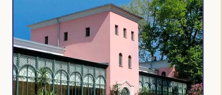 Ein rosafarbenes Gebäude mit Bogenfenstern und einem großen Gewächshaus. Vor dem Gebäude stehen mehrere Topfpflanzen.