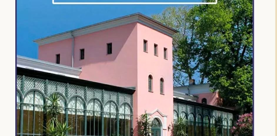 A pink building with arched windows and a large greenhouse. Several potted plants are in front of the building.
