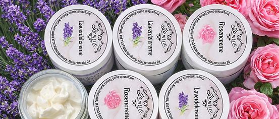 A collection of white tubs of lavender and rose cream, arranged on a bed of lavender and pink roses. Each tub is labeled with the product name and brand.