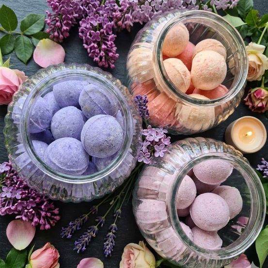 Vier Glasdosen mit Badebomben, zwei lila und zwei rosa, umgeben von Flieder, Lavendel, Rosen und grünen Blättern.