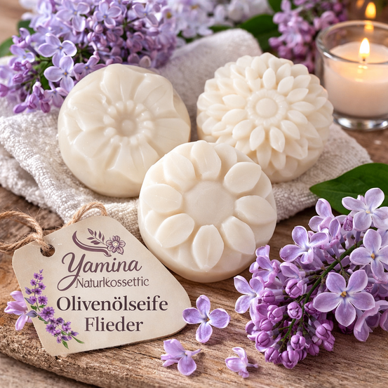 Drei handgemachte Seifenstücke mit Blumendesigns liegen auf einem weißen Handtuch mit lila Flieder. Eine brennende Kerze und Blätter sind in der Nähe. Ein Schild zeigt 'Yamina Naturkosmetik Olivenoelseife Flieder'.