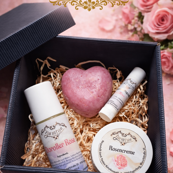 Eine schwarze Box mit einem herzförmigen Badeseife und zwei kosmetischen Produkten. Die Box ist mit Stroh gefüllt und hat einen dekorativen Blumenhintergrund.