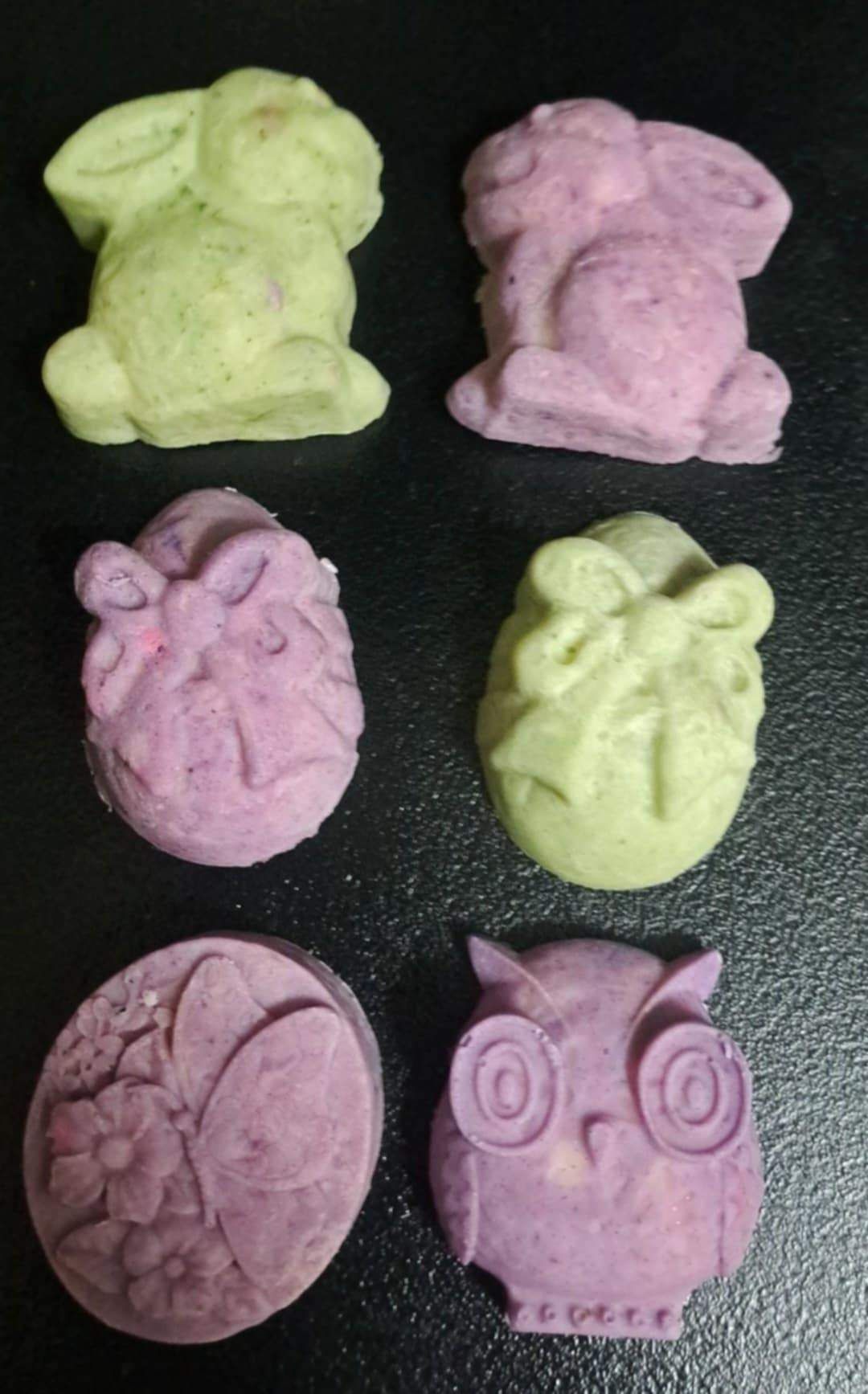 Sechs verschiedenfarbige und geformte Seifen liegen auf einer schwarzen Oberfläche. Die Farben sind lila, grün und pink.