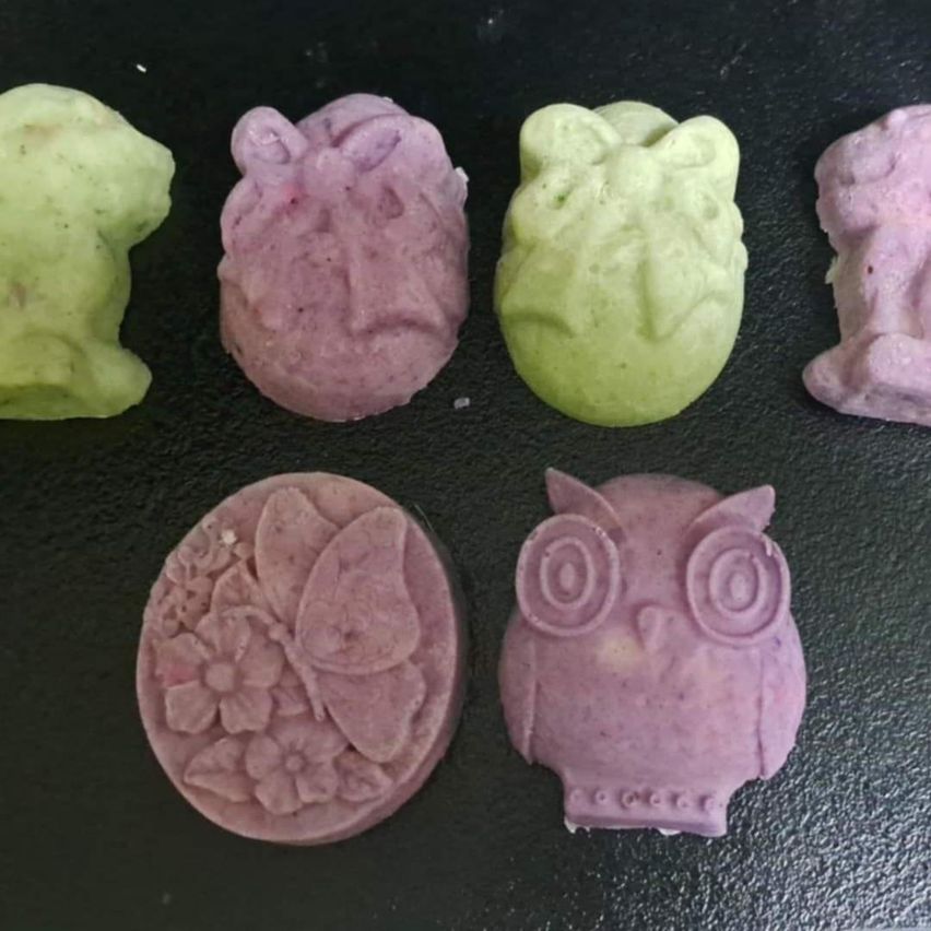 Sechs bunte Seifen in Form von Tieren und Insekten auf einer schwarzen Oberfläche. Eine ist eine Eule, eine andere ein Schmetterling, und andere sind ein Frosch, ein Vogel und eine Katze.