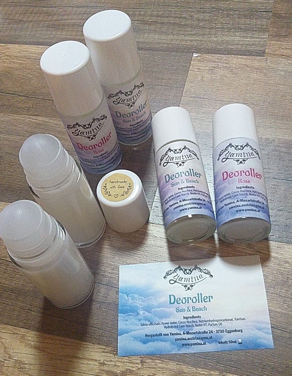 Mehrere Glas- und Plastikflaschen mit Kappen liegen auf einem Holztisch. Die Etiketten auf den Flaschen lesen 'Deorolller', 'Sonne & Strand' und 'Rose'. Es gibt eine Visitenkarte mit einem Design und dem Text 'Deorolller Sonne & Strand'.