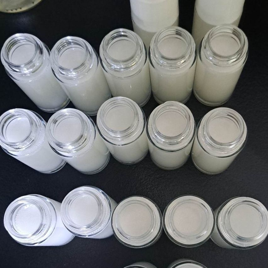 Mehrere Glasflaschen mit weißen, undurchsichtigen Inhalten sind auf einer schwarzen Oberfläche aufgereiht. Die Flaschen haben silberne Deckel und der Inhalt scheint eine glatte, möglicherweise flüssige oder halbfeste Substanz zu sein.